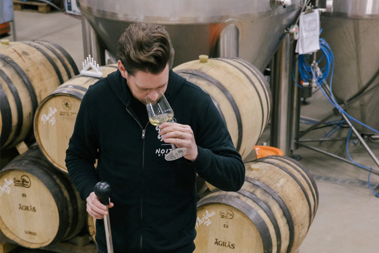 Mies haistelee viininäytettä viinitynnyreiden keskellä.
